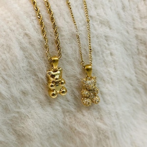 May include: Two gold-colored necklaces with teddy bear pendants. One pendant is a solid gold bear, and the other is encrusted with small, sparkling stones. The chains have different designs, one a rope chain and the other a beaded chain.