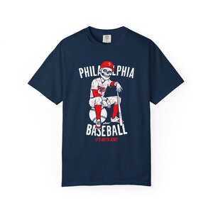 May include: Navy blue t-shirt featuring a baseball-themed graphic. The design includes a skeleton in a baseball uniform, a baseball bat, and the words "PHILADELPHIA BASEBALL" and "IT'S OUTTA HERE!" in white and red.