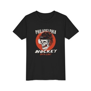 May include: Black t-shirt featuring a graphic of a skull wearing a hockey helmet, set against a red circle. The text "PHILADELPHIA" arches above the skull, with "HOCKEY UNTIL I DIE!" below.