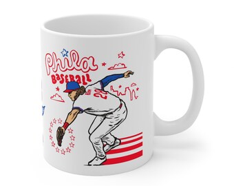 May include: White ceramic mug featuring a baseball player illustration. The design includes the words "Phila Baseball" in red and blue, along with stars and a baseball player in action. The mug has a curved handle.