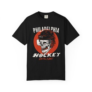 May include: Black t-shirt with a graphic of a skull wearing a hockey helmet and the words "PHILADELPHIA HOCKEY UNTIL I DIE!" in white and red. The skull is centered on a red circle.