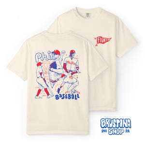 Può includere: Una t-shirt a maniche corte color crema con una grafica rossa e blu sul retro che raffigura tre giocatori di baseball in azione, il testo "Phils", "2026" e "BASEBALL". Sul petto è presente un piccolo gagliardetto rosso con "FIGHTINS".