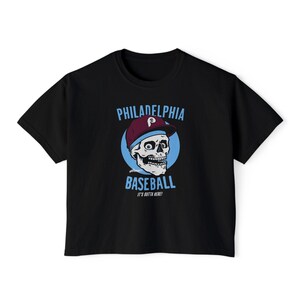 May include: A black short-sleeve t-shirt featuring a graphic of a skull wearing a maroon and blue baseball cap. The text reads PHILADELPHIA BASEBALL and IT'S OUTTA HERE! in light blue letters against a circular blue background on the chest.