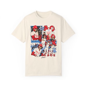 May include: A cream-coloured t-shirt featuring a graphic print of baseball players in red and blue uniforms. The graphic includes the text "A-O-K" and "Stott Shot".