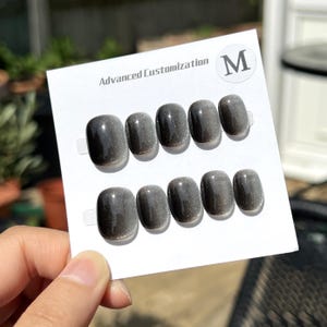 Peut inclure: Un ensemble de faux ongles gris avec un effet scintillant. Les ongles sont de forme ovale et disposés sur une carte blanche. La carte porte le texte "Advanced Customization" et un cercle avec la lettre "M".