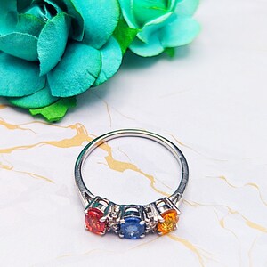 Könnte beinhalten: Ein silberner Ring mit drei bunten Edelsteinen. Der Ring hat einen runden orangen Stein, einen runden blauen Stein und einen runden gelben Stein. Die Steine sind in einer silbernen Fassung gefasst.