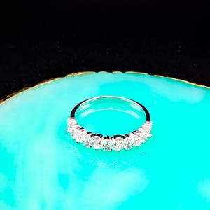 Könnte beinhalten: Ein silberner Ring mit mehreren klaren, quadratisch geschliffenen Steinen in einer Reihe. Der Ring wird auf einer leuchtend türkisfarbenen Oberfläche mit goldfarbenem Rand vor schwarzem Hintergrund präsentiert. Der Ring ist ein Schmuckstück.