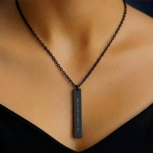 May include: A black necklace with a rectangular pendant. The pendant has raised dots in a vertical line, likely representing Braille. The chain and pendant are a matching dark color, creating a sleek, minimalist design. The necklace is worn against a dark background.