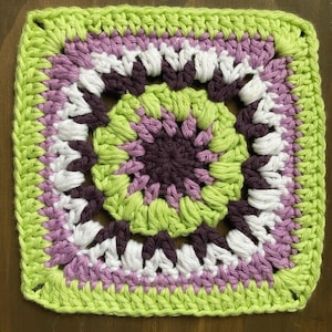 Op de afbeelding: Een vierkant gehaakt granny square met een cirkelvormig patroon in het midden. Het ontwerp gebruikt concentrische ringen van garen in tinten limoengroen, lavendel, donkerpaars en wit, gewerkt in een herhalend steekpatroon dat overgaat van cirkel naar vierkant.