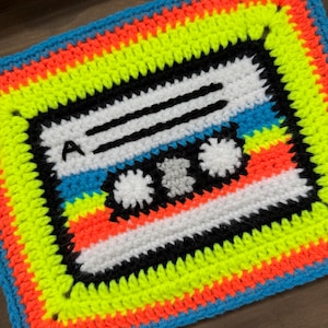 Puede incluir: Una pieza cuadrada de ganchillo con un diseño de cinta de casete. El centro es blanco con líneas horizontales negras y dos carretes circulares blancos. El borde consta de filas concéntricas de puntos de hilo amarillo neón, naranja y azul.