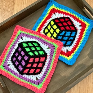 May include: Two square crochet coasters featuring a central cube design. One coaster has a blue, red, yellow, and white border with a multi-colored cube. The other has a pink, purple, green, and white border with a green, purple, and pink cube.