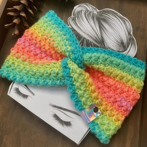 May include: A handmade, rainbow-colored crochet headband. The headband features a vibrant gradient of colors, including pink, orange, yellow, green, and blue. A small, iridescent button and strap detail is visible on the side. The headband is displayed on a wooden surface.