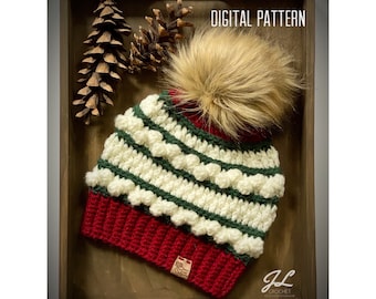 May include: A white and green crocheted beanie with a red brim and a faux fur pom pom. The beanie has a textured design with raised bumps. The beanie is displayed in a wooden tray with pine cones.