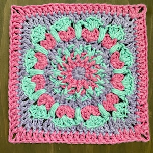 Op de afbeelding: Een vierkant haakmotief met een cirkelvormig bloemenpatroon in het midden. Het ontwerp gebruikt concentrische lagen garen in tinten felroze, lichtlavendel en mintgroen, die overgaan in een effen roze rand aan de buitenkant.