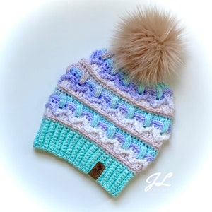 Op de afbeelding: Een gebreide muts met een kleurrijk gestreept patroon in tinten van paars, roze, wit en turquoise. De muts heeft een pluche bruine pompon bovenop en een bruin leren label met een hartje. De muts is gemaakt met een textuur-steek en heeft een slouchy pasvorm.