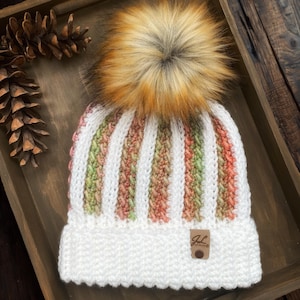 Könnte beinhalten: Gestrickte Mütze mit Kunstfell-Bommel. Die Mütze hat weiße, grüne und pfirsichfarbene Streifen. Ein kleines Lederetikett mit den Initialen "JL" ist am Rand angenäht. Die Mütze wird auf einer Holzoberfläche präsentiert.