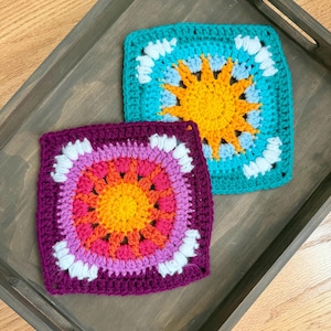 Op de afbeelding: Twee vierkante gehaakte onderzetters met een zonnestraalpatroon. De ene onderzetter heeft een felgeel midden met oranje en turquoise garen, terwijl de andere een geel midden heeft met oranje, roze en donkerpaars garen, beide met witte accenten.