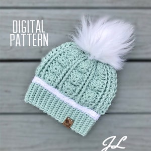 May include: A light green crochet beanie with a white pom pom and a white crocheted band. The beanie has a brown label with the text "Crochet Creations" on it. The text "DIGITAL PATTERN" is in the top left corner of the image. The text "JL CROCHET" is in the bottom right corner of the image.