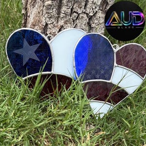 May include: Two heart-shaped stained glass suncatchers featuring a patriotic design with red, white, and blue segments. One heart displays a prominent star on a blue background, while the other features a striped pattern. Both have metal solder lines.