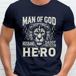 May include: Navy blue t-shirt with a graphic of a lion's head and the words "MAN OF GOD, HUSBAND, DADDY, PROTECTOR, HERO" in white. The design includes lightning bolts and mountain silhouettes.