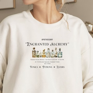 May include: White sweatshirt with a graphic print of vintage apothecary bottles and the text "APOTHECARY" "ENCHANTED ALCHEMY" "OPENING HOURS MONDAY SATURDAY 10:30 AM 07:30 PM AT 73 WICKED SQUARE DAEMON TOWN EST 1602 TONICS POTIONS ELIXIRS"