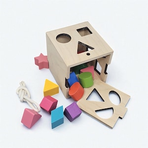 May include: A wooden shape sorter toy with a natural finish. The box has cut-out shapes and is tilted, with colorful geometric blocks spilling out. A pink star and a rope are also present.