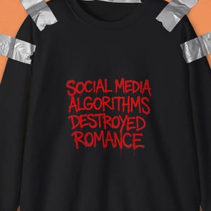 Puede incluir: Una sudadera negra de cuello redondo con el texto rojo SOCIAL MEDIA ALGORITHMS DESTROYED ROMANCE impreso en una fuente de estilo grafiti que gotea en el pecho. La prenda está fijada a una superficie naranja con trozos de cinta adhesiva gris.