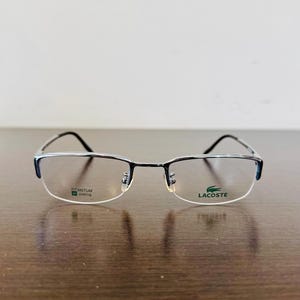 Peut inclure: Lunettes rectangulaires avec une monture noire et argentée. Les verres transparents sont encadrés par une monture avec le texte "TITANIUM IP-plating" sur le verre gauche et le logo Lacoste sur le verre droit.