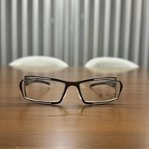 Może przedstawiać: Okulary z ciemnobrązową oprawką i prostokątnymi soczewkami. Okulary mają unikalny design z cienkim białym paskiem wzdłuż krawędzi. Tekst "parasite" jest widoczny na prawej soczewce.