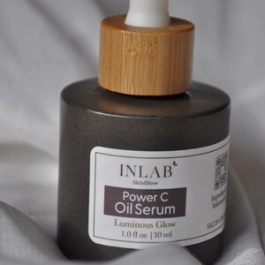 May include: A dark grey glass bottle with a bamboo lid and a white dropper top. The bottle contains a serum called "Power C Oil Serum" by Inlab SkinGlow. The label also says "Luminous Glow" and "1.0 fl oz | 30 ml".