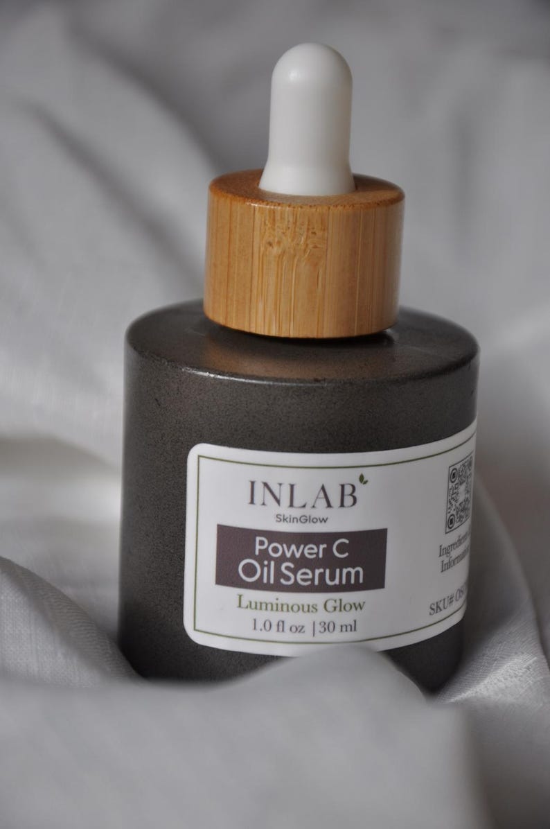 May include: A dark grey glass bottle with a bamboo lid and a white dropper top. The bottle contains a serum called "Power C Oil Serum" by Inlab SkinGlow. The label also says "Luminous Glow" and "1.0 fl oz | 30 ml".