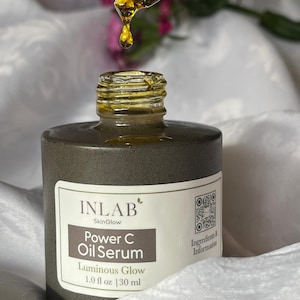 May include: A brown glass bottle with a white label that reads "INLAB SkinGlow Power C Oil Serum Luminous Glow 1.0 fl oz | 30 ml". The bottle is on a white surface and a dropper with a yellow liquid is above the bottle.