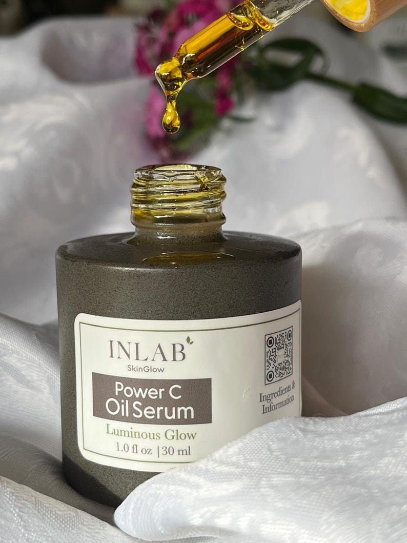 May include: A brown glass bottle with a white label that reads "INLAB SkinGlow Power C Oil Serum Luminous Glow 1.0 fl oz | 30 ml". The bottle is on a white surface and a dropper with a yellow liquid is above the bottle.