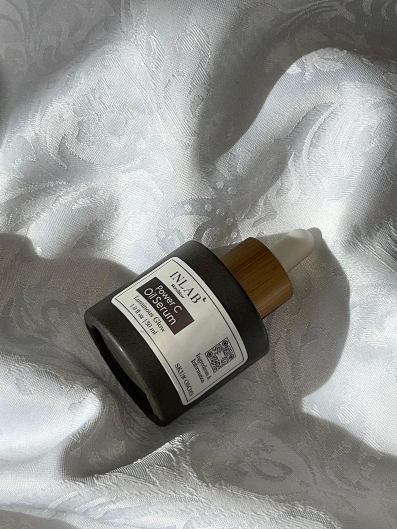 May include: A dark grey glass bottle with a bamboo dropper top. The bottle is labelled "INLAB" "Power C Oil Serum" "Luminous Glow" "1.0 fl oz (30 ml)" and "SKU# OSO".