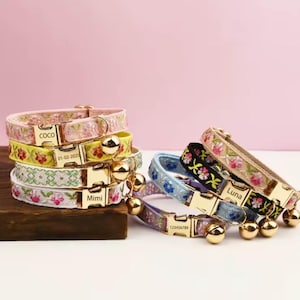 Puede incluir: Una colección de collares bordados para gatos en varios colores, incluyendo rosa, amarillo y azul. Cada collar presenta una hebilla dorada, una pequeña campana y texto personalizado. Los collares están apilados sobre un bloque de madera.