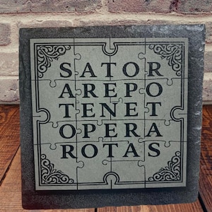 May include: A square, grey stone puzzle featuring the Latin palindrome "SATOR AREPO TENET OPERA ROTAS". The puzzle pieces are arranged in a grid, with decorative corner flourishes. The background shows a brick wall and wooden surface.