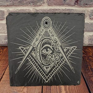 May include: A black slate plaque with a detailed, engraved Masonic symbol. The symbol features a compass, square, and the all-seeing eye, with radiating lines. The design is in a lighter color, contrasting against the dark slate.