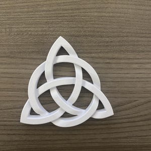 May include: A white, three-pointed Celtic knot symbol, also known as a triquetra, is displayed against a brown wooden surface. The knot is a continuous line, symbolizing interconnectedness and eternity. The design is a popular symbol in Celtic art and spirituality.