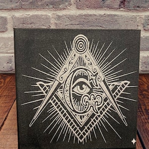 Peut inclure: Toile noire avec un symbole maçonnique blanc représentant un compas, une équerre et un œil omniscient. L'œuvre d'art est posée sur un mur de briques et une surface en bois. Une étoile est visible en bas à droite.