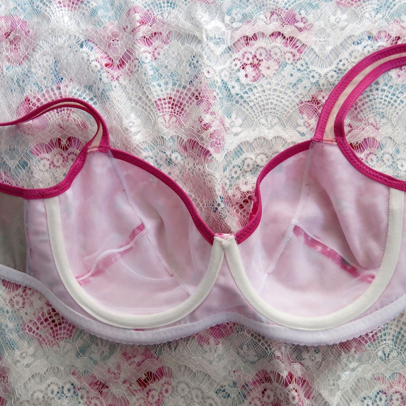 May include: A pink bra with white trim and a pink lace pattern. The bra is on a white lace background.
