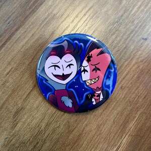 May include: A round button with a black and red cartoon character design. The characters are smiling and have a starry blue background.
