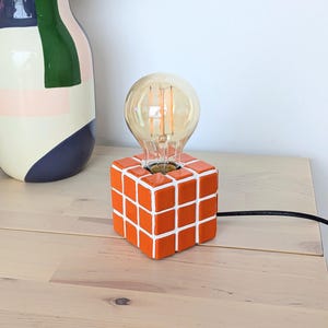 Op de afbeelding: Oranje en witte kubusvormige lamp met een grote, ronde Edison-stijl gloeilamp. De lamp staat op een houten oppervlak. Een zwart netsnoer loopt van de lamp. Een decoratieve vaas met een groen, blauw en roze ontwerp staat op de achtergrond.