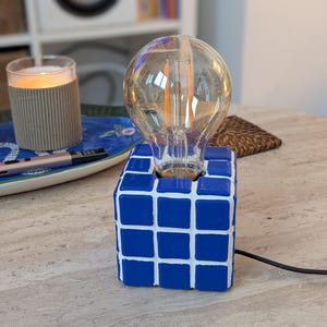 Op de afbeelding: Een unieke tafellamp met een grote, heldere Edison-lamp op een blauw-witte kubusvormige basis. De lamp heeft een zwart snoer en staat op een lichtgekleurd oppervlak. De lamp is aan en geeft een warme gloed.