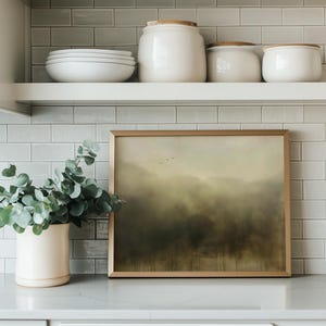 May include: A framed landscape painting with a muted green and brown color scheme hangs on a white tiled wall above a white shelf. The painting depicts a misty forest scene with a hint of blue sky in the distance. A potted eucalyptus plant sits on the counter below the shelf.