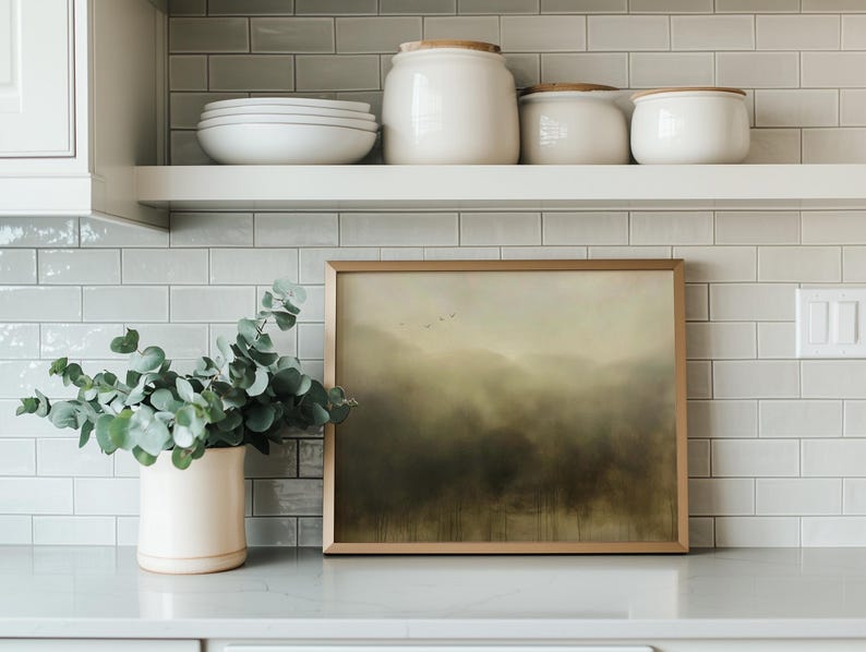 May include: A framed landscape painting with a muted green and brown color scheme hangs on a white tiled wall above a white shelf. The painting depicts a misty forest scene with a hint of blue sky in the distance. A potted eucalyptus plant sits on the counter below the shelf.