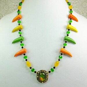 May include: A colorful necklace with green, yellow, and orange beads and a large green gemstone pendant. The necklace is made of glass beads and has a gold-tone finish.