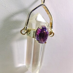 May include: A clear quartz crystal pendant with a silver-colored metal setting. The pendant features a faceted, pointed top and a round, purple amethyst stone. The crystal hangs from a silver chain.