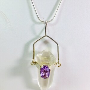 May include: A silver pendant necklace featuring a clear quartz crystal with a faceted top and a small, oval, purple amethyst. The pendant is suspended from a delicate silver chain.