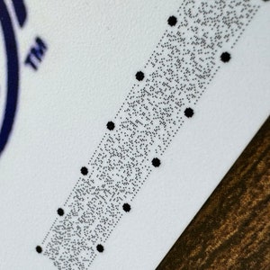 May include: A close-up of a white card with a black and white braille pattern. The card has a blue and white logo in the top left corner and the word "TM" in the top right corner.