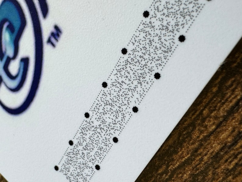 May include: A close-up of a white card with a black and white braille pattern. The card has a blue and white logo in the top left corner and the word "TM" in the top right corner.